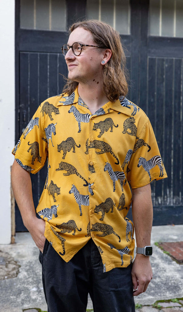 white male model with long hair and glasses wearing Golden Zebra Short Sleeve Rayon Shirt paired with black jeans. The shirt is a yellow gold colour with an all over print of zebras and big cats. The model is stood outside in Hove posing toward the camera with one hand in his jeans pocket.