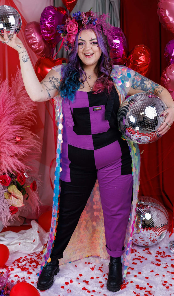 Luisa, a white femme model with purple, blue and pink in their hair is stood in a studio in Hove with a red backdrop with heart shaped balloons and pink feathers and rose petals on the floor. They are wearing Purple and Black Mix n Match Two Tone Stretch Twill Dungarees with a sequin shawl and floral headdress. Luisa is posing holding disco balls and facing the camera. 