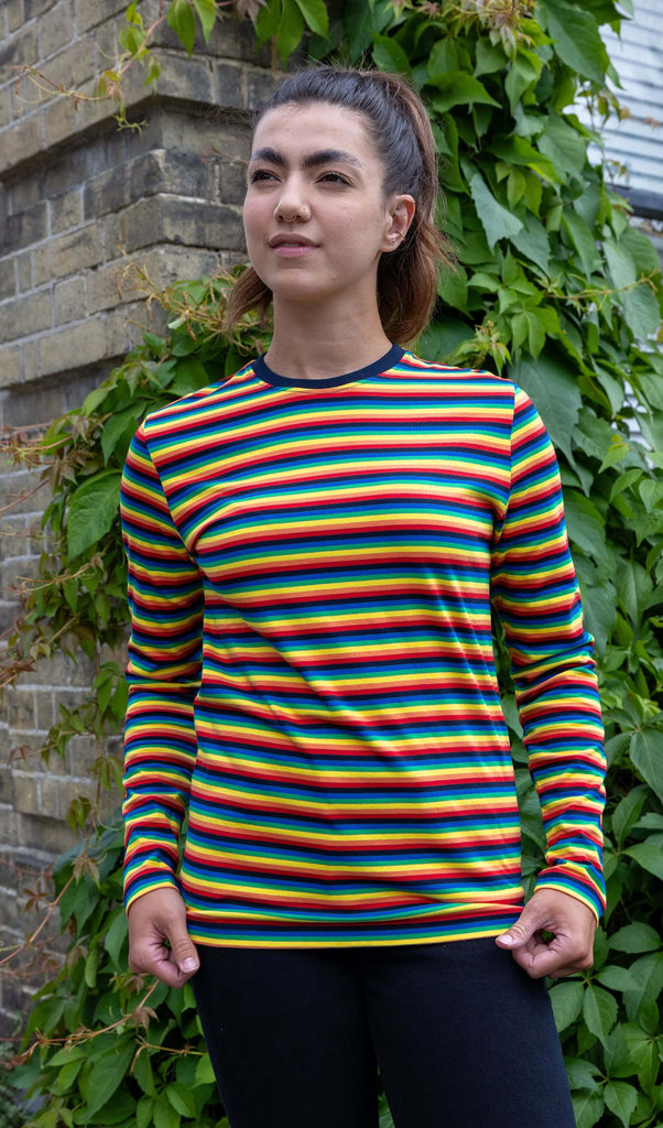 Sophie is wearing Retro Rainbow Brights Repeat Striped Long Sleeved T Shirt with black trousers. She is stood outside in Hove in front of a brick wall with greenery and is posing facing the camera looking off into the distance while holding the bottom of the t shirt.