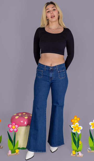 Shannon is wearing Stonewash Blue Stretch Denim Bell Bottom 90's Wide Leg Hippy Flares with a black cropped long sleeve top and white boots. She is stood in front of a grey backdrop with a toad stool and flower decorations next to her. She is posing toward the camera with her hands behind her back.