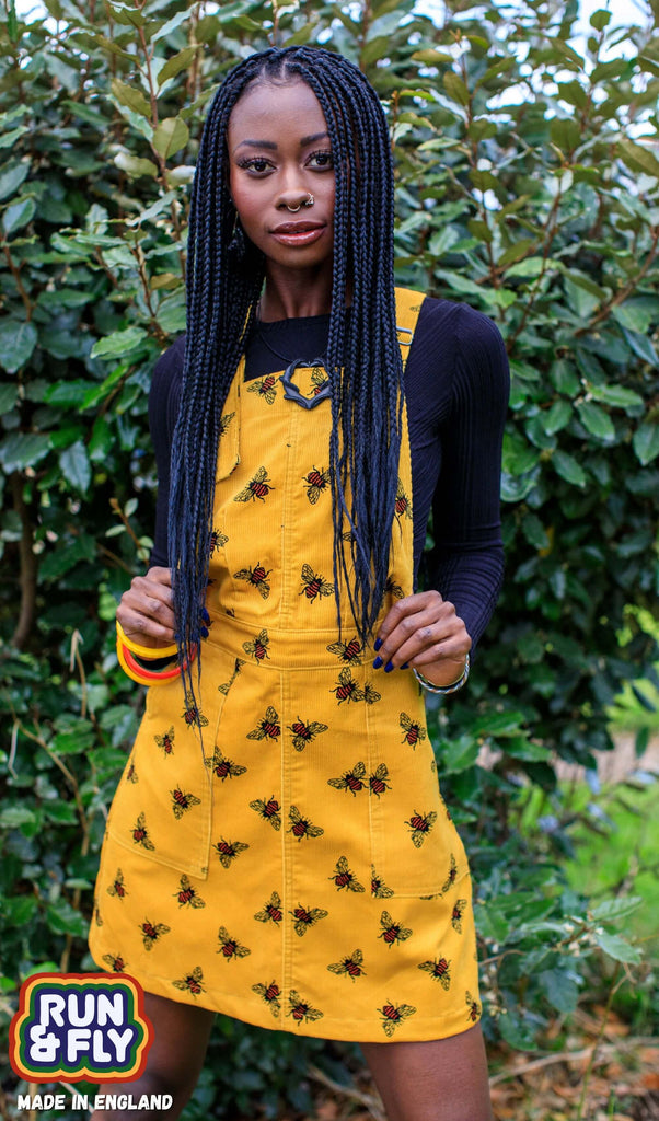 Carella is stood in front of a bush wearing the mustard corduroy the bees kneez bee print pinafore dress with a long sleeve black tshirt underneath. They are facing the camera and holding onto the bottom of the pinafore bib whilst smising. Photo is cropped from the knees up.