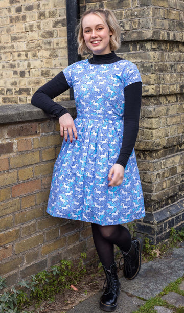 Femme model with short blonde hair is stood outside by a brick wall wearing Unicorn Tea Dress with a long sleeve black turtle neck underneath and black tights and boots. The blue dress has an all over unicorn print. The model is stood leaning against a wall smiling at the camera with one leg bent. 