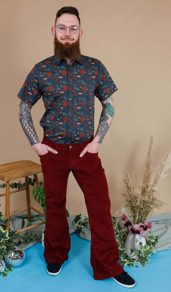 Dec, a white male with a beard and glasses, is stood in a photography studio in Hove in front of a beige backdrop wearing Tawney Port Corduroy Bell Bottom Flares with Retro Old School Tattoo Print Short Sleeve Shirt. Dec is smiling at the camera with his hands in the trouser pockets. 