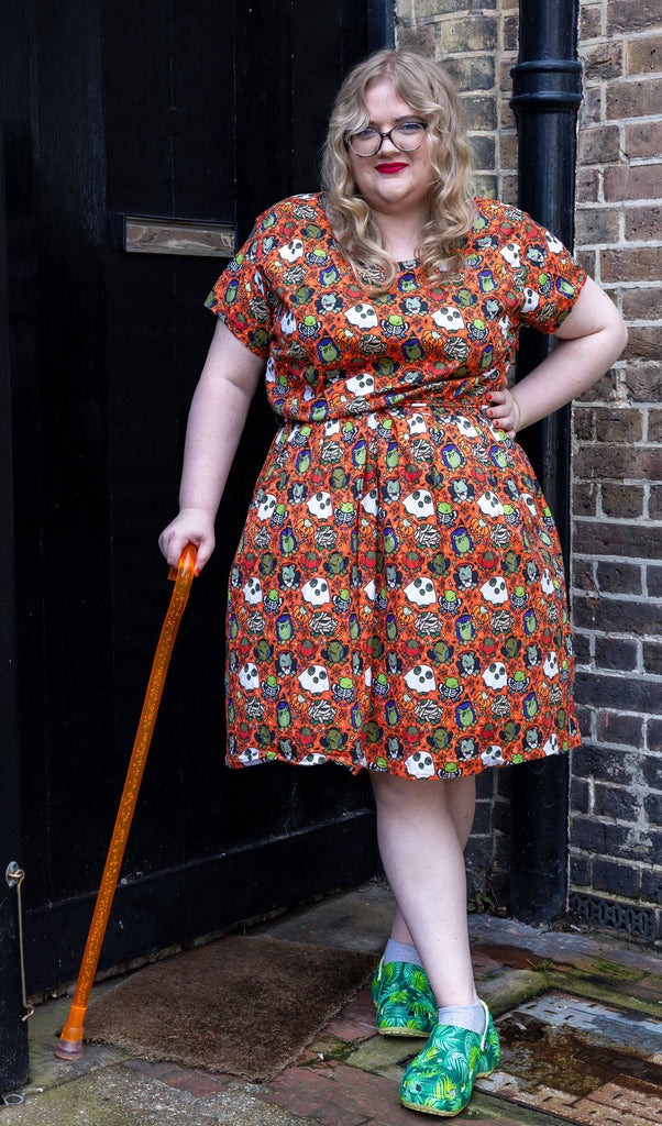 a white femme model with red lipstick wearing Halloween: Party Frogs Stretch Belted Tea Dress with Pockets. The orange dress has an all over print of cartoon frogs in various Halloween costumes such as ghosts and zombies. The model is stood outside in Hove smiling towards the camera with one hand on their hip and other holding an orange walking aid.