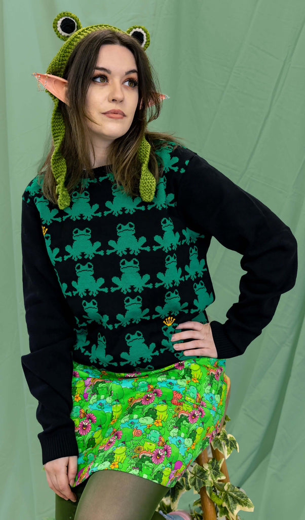 Alice is sat in front of a draped sheet background wearing the frog prince jumper with the mushroom babes frogs stretch twill pinafore, green tights, frog hat and elf ears. She is leaning slightly forward with one hand on her hip and smiling looking right. The jumper is a black base with repeating green frogs (two have gold crowns), the arms and back are plain black.