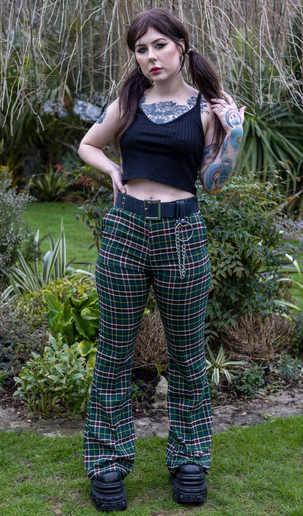 Florence is stood outside in a garden in Hove wearing Green Tartan Plaid Bell Bottom Slacks paired with black top, black belt with chain, and chunky black boots. She is facing the camera and posing with one hand in her hand and other on her hip. 