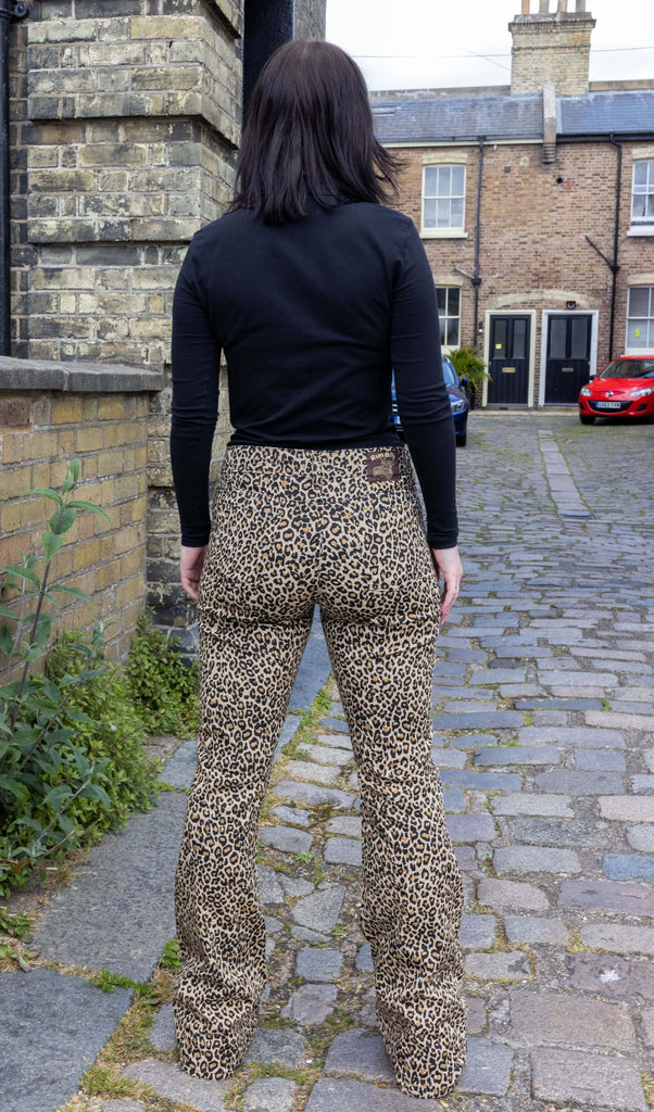 Model with dark hair wearing Leopard Print Bell Bottom Stretch Super Flares, paired with a long sleeve black turtleneck top and chunky black boots. The flares are a natural leopard print pattern and colour. Model is posing with back to camera.