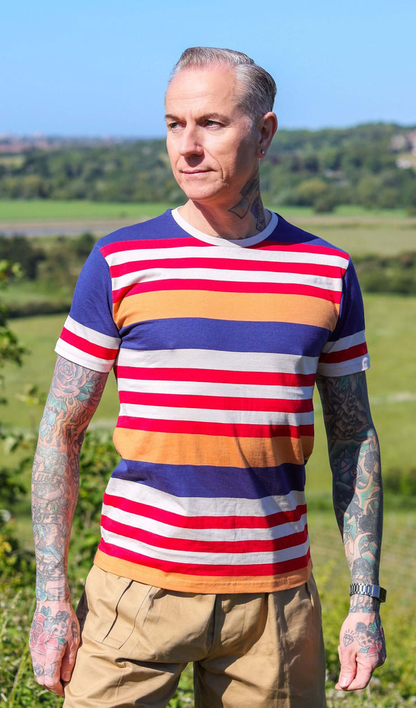 Jim is stood on a grass hill wearing the retro multi striped nugget tshit with beige cargo trousers. They are heavily tattooed with swept back grey hair. They are facing forward posing with both arms resting by their side and looking off to the left. Photo is cropped from the hips up.