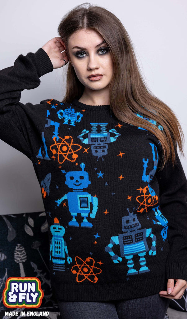 Charlotte is stood in front of a grey sofa wearing the robot jumper with black stone wash flares. She is facing forward with one hand in her hair and other resting by her side and is posing at the camera. Photo is cropped from the thighs up. The robot jumper is a black base featuring multiple blue robots with orange, blue stars and chemical symbols with plain black arms and back.
