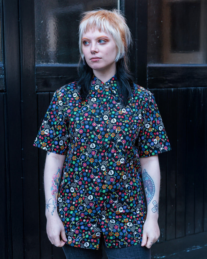 Faeryn is stood outside wearing the halloween: boogie bones short sleeve shirt with black stonewash flares. They are facing forward looking off to the left with both arms resting by their side. The photo is cropped from the thighs up. Boogie bones print is a black base with multicoloured flowers and dancing smiling cartoon skeletons all over.
