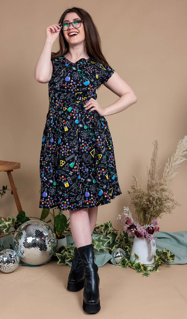 Charlotte is stood in front of a beige backdrop with various plants and disco balls behind her. She is wearing School of Science Belted Tea Dress with Pockets with black boots. The dress has a black background colour with an all over print of various chemistry equipment in white, yellow, green, red, yellow and purple. Charlotte is smiling and holding onto her glasses with her ankles crossed and hand on her hip.