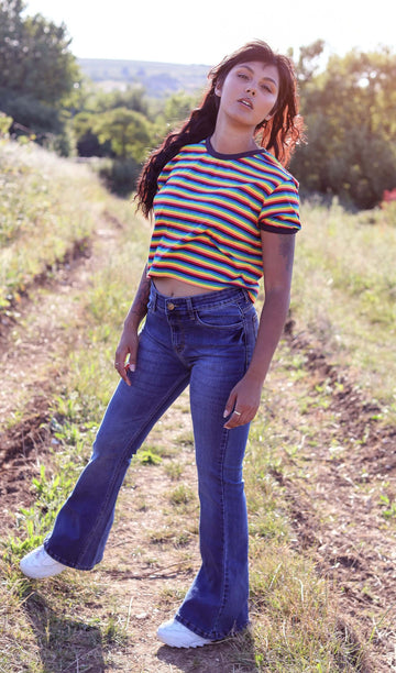 Meera is stood in a grassy field wearing the stone wash blue distress stretch denim rock n roll bell bottom flares with a retro rainbow short sleeve tshirt and white trainers. She is facing forward posing on one leg and leaning slightly back whilst looking to camera.