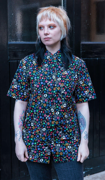 Faeryn is stood outside wearing the halloween: boogie bones short sleeve shirt with black stonewash flares. They are facing forward looking off to the left with both arms resting by their side. The photo is cropped from the thighs up. Boogie bones print is a black base with multicoloured flowers and dancing smiling cartoon skeletons all over.
