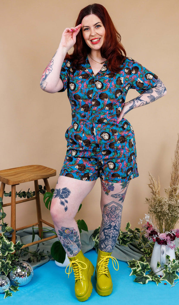 Tattooed model with red hair wearing Hedgehog Stretch Playsuit paired with lime green chunky boots. The playsuit is a blue colour with an all over print of brown hedgehogs and green, pink and orange leaves. The collared playsuit is buttoned up, with the top button left undone. Model is posing with one hand on hip and other hand in her hair.