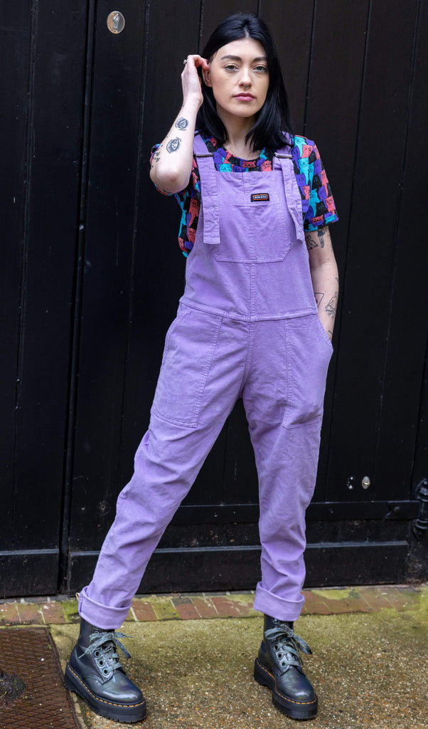 white femme model with black hair and tattoos is stood outside in Hove in front of a black door wearing Lavender Stretch Corduroy Dungarees paired with Cat Chorus Short Sleeve Tee and silver boots. The model is posing facing the camera with one hand in the dungaree pocket and other tucking her hair behind her ear.  