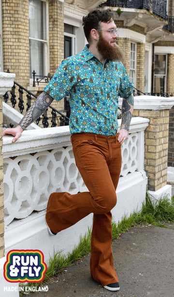 Dec is stood outside a town house in Hove wearing the Tobacco Corduroy Bell Bottom Super Flares Trousers with Bunny Meadow Short Sleeve Shirt. He is leaning against a wall with one leg bent and his hand on his hip. He is smiling looking off into the distance.