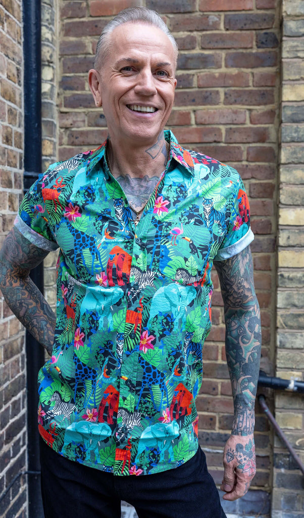 James is wearing Jungle Stretch Short Sleeve Shirt, paired with black trousers. The collared shirt is a forest green colour with all over print of various jungle animals and plants in black, white, red, pink, yellow and blue. James is smiling at the camera with one hand on his hip.