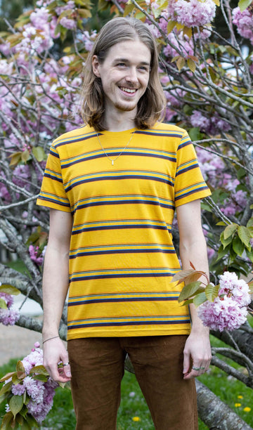 Jack is stood in front of a blossom tree wearing the gold sunset stripe retro tshirt with tobacco corduroy flares. He has mid length blonde hair and is smiling facing towards the camera with both hands resting by his sides. Photo is cropped from the knees up.