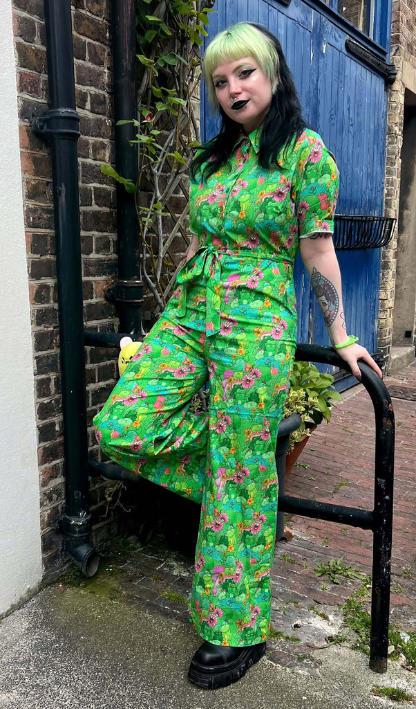 Tattooed model with green and black hair and dark makeup wearing Run & Fly x The Mushroom Babes Frogs Stretch Jumpsuit, paired with black chunky boots and frog bangle. The jumpsuit is a bright green colour with an all over pattern of green frogs and lily pads, blue puddles, pink and orange flowers and mushrooms and various insects. The collared jumpsuit is buttoned up to the top and matching fabric belt is tied in a bow round front of waist. Model is leaning against pole and smiling at camera.