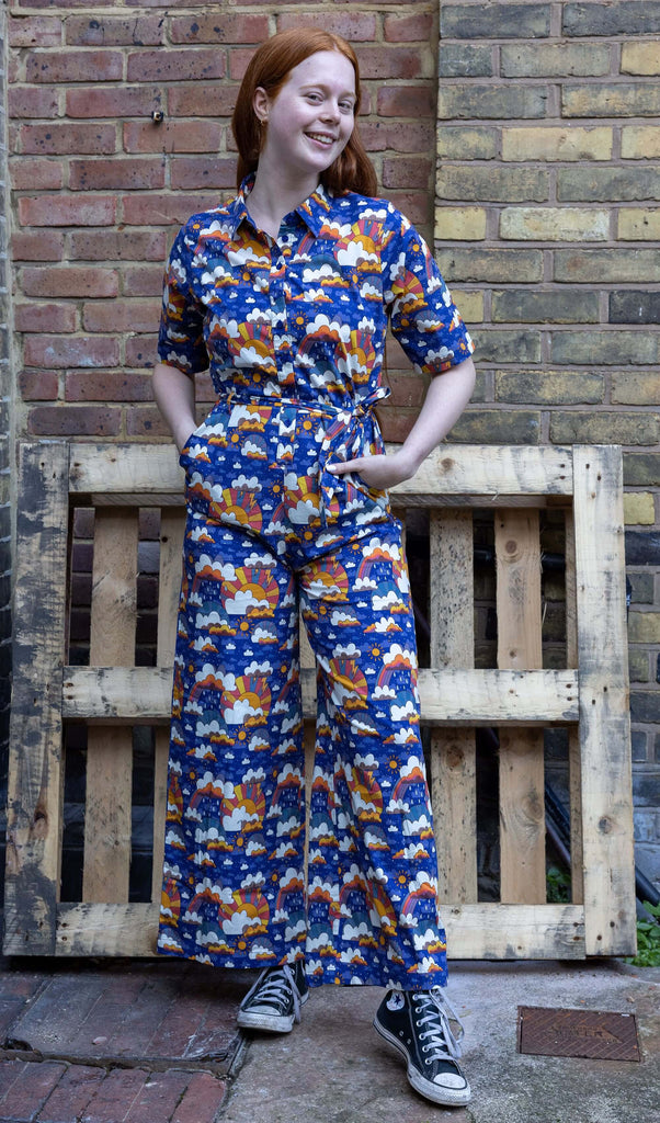 Sophie stood outside in a brick courtyard wearing the rainy days jumpsuit with black trainers, design by Jen James Design. The jumpsuit is short sleeved, collared, button down with waist tie belt with wide legs. The print is a dark blue base with yellow, orange, yellow and dusky purple clouds with raindrops, sunshines, sunbursts and rainbows. Sophie is stood facing forward smiling with both hands in the pockets and one leg pointed forward to highlight the wide leg shape.