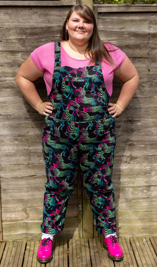 Natalie is wearing Rainbow Zebra Stretch Twill Dungarees with a pink t-shirt underneath and pink boots. The black dungarees have an all over colourful zebra print. She is stood outside in front of a wooden fence facing the camera smiling with her hands on her hips. 