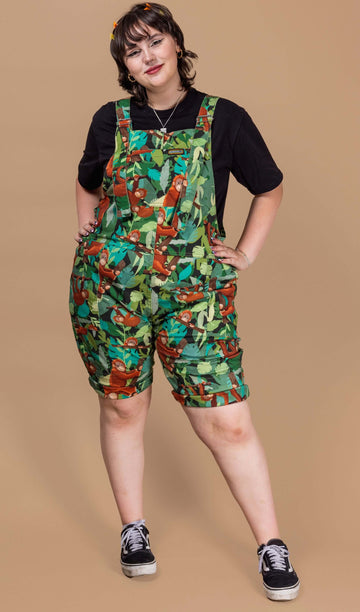 A white femme model with short brunette hair is wearing Orangutan Stretch Twill Dungaree Shorts with a black short sleeve t-shirt underneath. The green dress has an all over orangutan print with leaves and branches. The model is stood in front of a beige backdrop posing towards the camera with their hands on their hips.