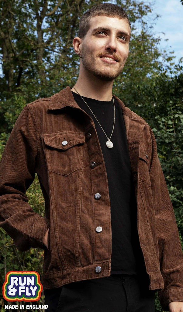 Jake is stood in a garden area smiling wearing the retro vintage cocoa brown cord unisex western jacket with a black tshirt underneath and black trousers. He is facing forward with one hand in the jacket pocket whilst looking up to the left. Photo is cropped from the hips up.