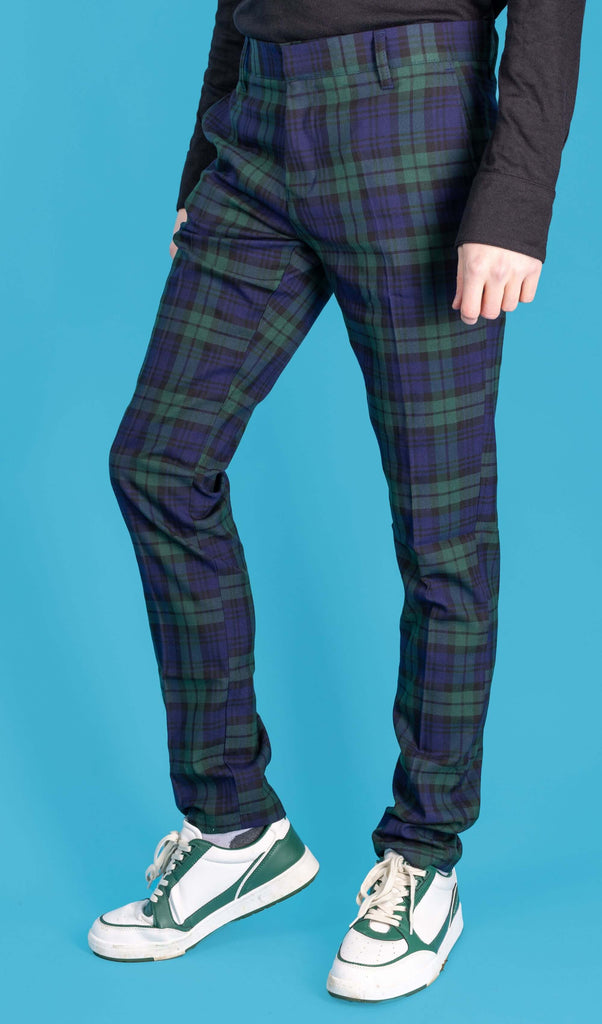 Jack is stood in front of a blue studio background wearing the retro mod stretch black watch tartan trousers with a long sleeve black tshirt and white trainers. They are facing towards the left with one leg bent forward to highlight the trouser shape. Photo is cropped from the waist down.