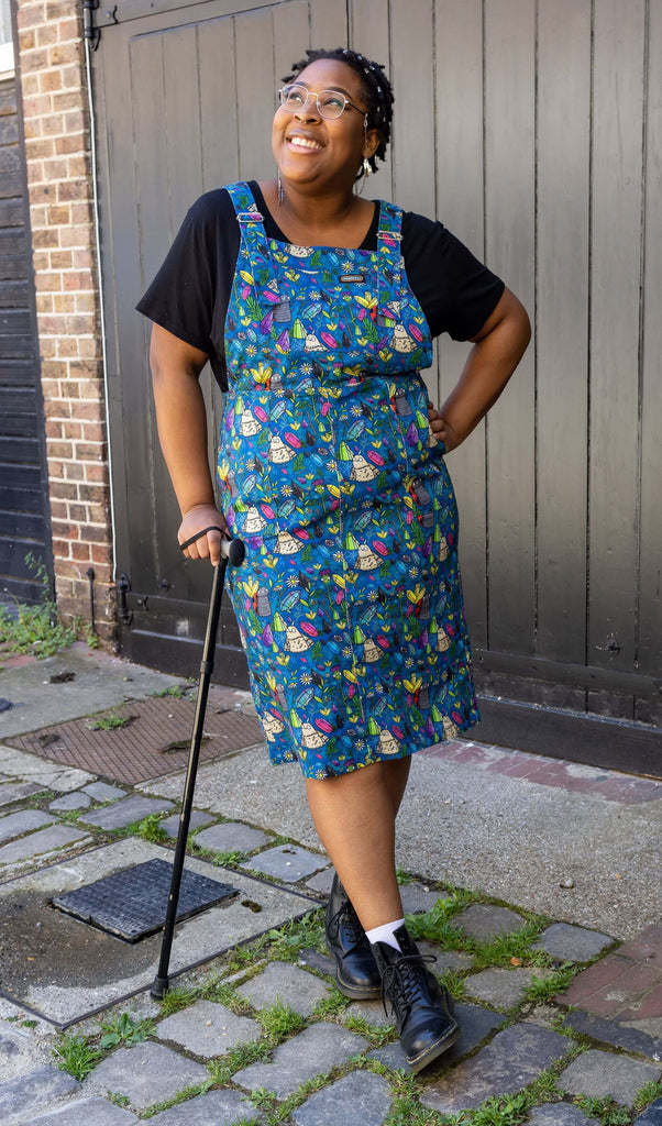 Han, a black femme model with silver glasses with a chain, is stood outside in Hove in a mews wearing Run & Fly x Katie Abey Crystal Critters Stretch Twill Long Pinafore Dress with a black t shirt underneath and black boots. The blue pinafore dress has an all over print of critters and crystals illustrated by Katie Abey. Han is smiling and is leaning on her black walking aid with her hand on her hip and one leg in front of the other. 