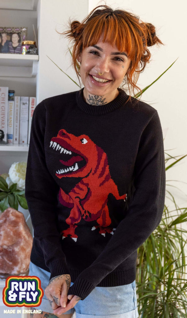 Amy is stood in a lounge wearing the t rex dino-roar jumper with blue denim shorts. They have short ginger hair and tattoos. They are facing forward posing with their hands clasped in front of them whilst smiling to camera. Photo is cropped from the hips up.