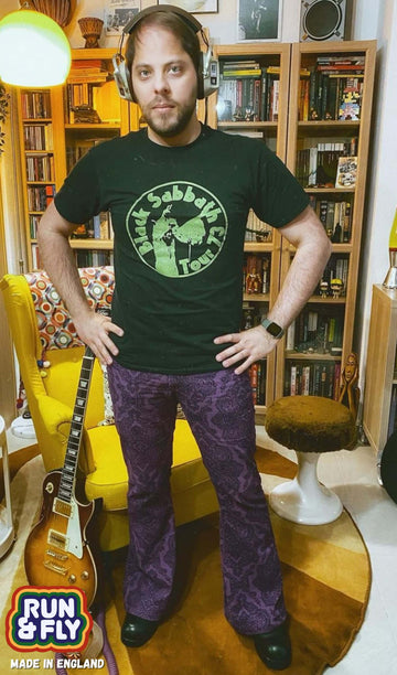 Stefano is stood in their 70s themed lounge next to their guitar and yellow chair wearing the purple grape Hendrix paisley corduroy retro bell bottom flares with a black sabbath tee and black boots. They are facing the camera posing with both hands on their hips and looking to the camera.