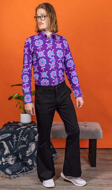 Jack is wearing Presley Black Stretch Cotton Twill Bell Bottom Trouser Slacks paired with Purple Retro Flowers Long Sleeve Shirt and white trainers. Jack is posing in a photography studio in Hove with an orange backdrop. He is posing looking off to one side with one leg bent and arms by his size. 