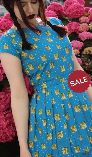 Alice is stood in front of a floral background wearing the kitsch kitty polka dot tea party dress. She is looking down to the left. Photo is cropped from the hips up.