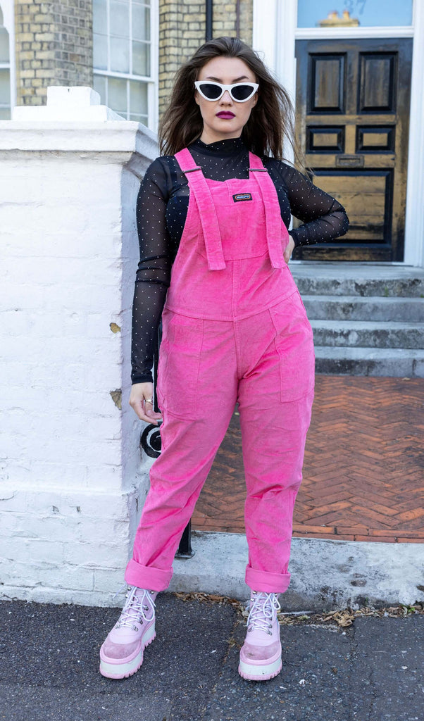Charlotte is wearing Pink Stretch Corduroy Dungarees, paired with a long sleeved black mesh top, white framed sunglasses and chunky pink boots. The dungarees are a bright pink all over colour with Run&Fly logo on the front bib. Charlotte is posing with hand on hip and bottoms of the legs of dungarees rolled up to reveal her boots.