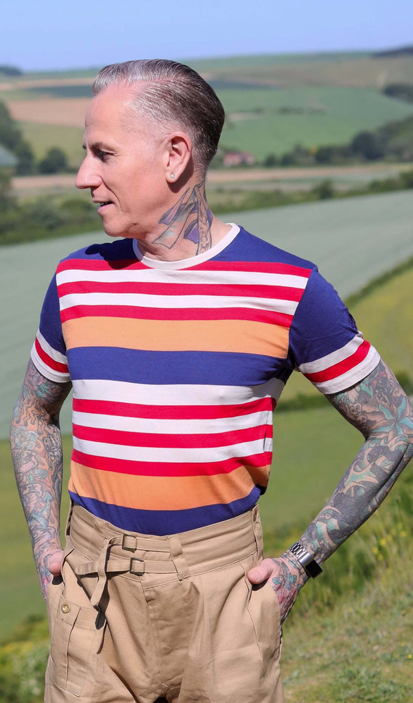 Jim is stood on a grass hill wearing the retro multi striped nugget tshit with beige cargo trousers. They are heavily tattooed with swept back grey hair. They are facing forward posing with both hands in their trouser pockets and looking off to the left smiling. Photo is cropped from the hips up.