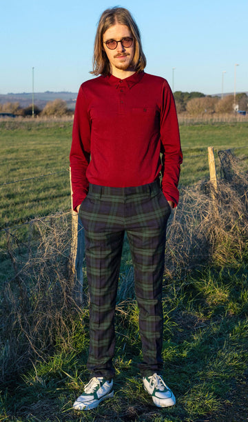 Jack is stood in front of a grass field wearing the retro mod stretch black watch tartan trousers with a long sleeve red shirt and white trainers. They are facing the camera posing with both hands in their trouser pockets and smiling to camera.