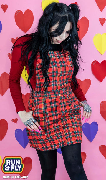 Rosie is stood in front of multicoloured hearts on a pink studio background wearing the red tartan pinafore dress with a long sleeve red tshirt and tights underneath. They have long black hair and tattoos. They are facing the camera posing with both hands resting in the front pinafore pockets and looking down to the right. Photo is cropped from the knees up.