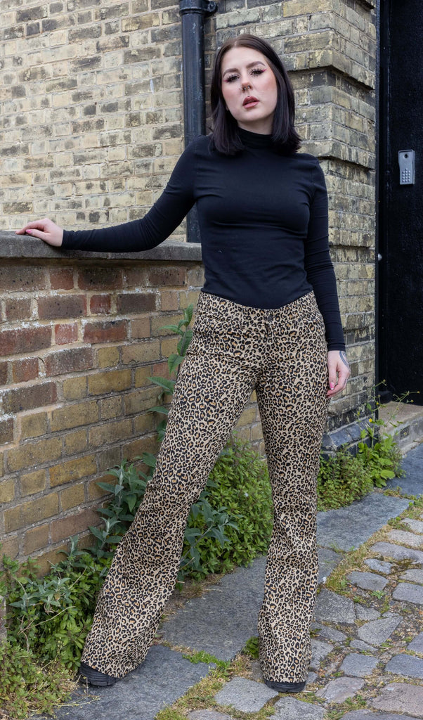 Model with dark hair wearing Leopard Print Bell Bottom Stretch Super Flares, paired with a long sleeve black turtleneck top and chunky black boots. The flares are a natural leopard print pattern and colour. Model is posing with one hand by her side and the other resting on a wall next to her.
