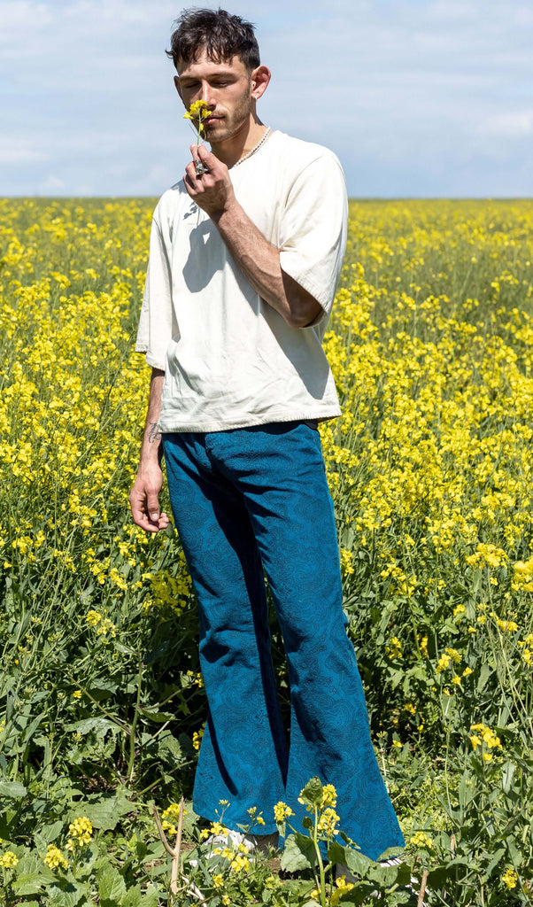  Zach is stood in a yellow flower field wearing the ink blue paisley corduroy bell bottom super flares with an oversized cream tshirt and white trainers. He is holding and smelling a yellow flower in front of his face.