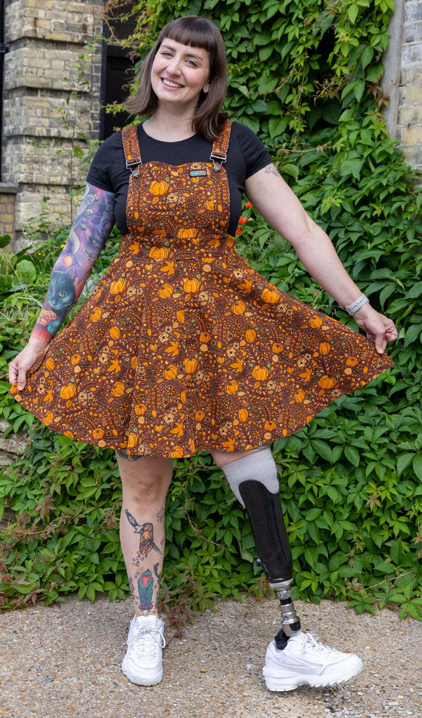 white tattooed model with brunette hair is stood outside in Hove wearing Halloween: Autumn Pumpkin Flared Pinafore Dress paired with a black tee underneath and white trainers. The rust coloured pinafore has an all over orange autumn pumpkin print. The model is smiling toward the camera holding the flared skirt of the dress. 