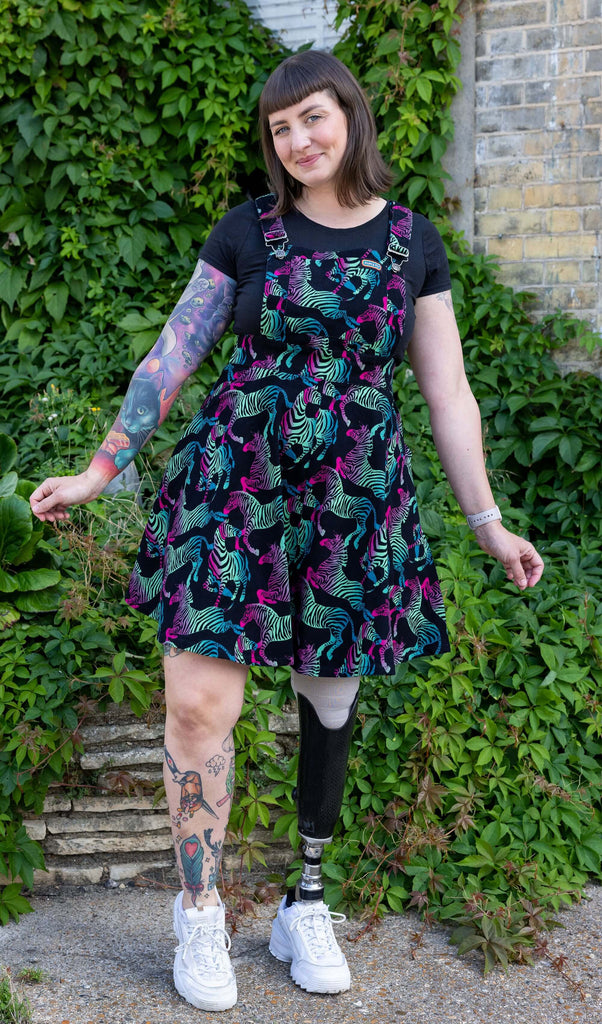 Laura, a white femme model with tattoos, is wearing Rainbow Zebra Flared Pinafore Dress with a black t-shirt underneath. The black pinafore dress has an all over colourful zebra print. Laura is stood outside in Hove in front of greenery facing the camera and smiling with her arms out to her sides and one leg slightly bent. 