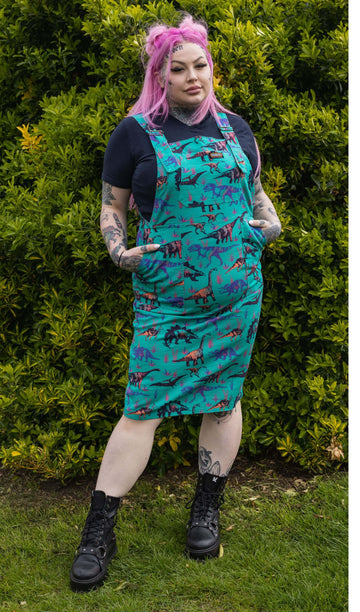 Zoe is stood in a garden area wearing the jade adventure dinosaur print stretch twill pinafore dress with a short sleeve black tshirt and black boots. She has long pink hair in half up space buns, facing the camera and posing with both hands in the pinafore pockets.