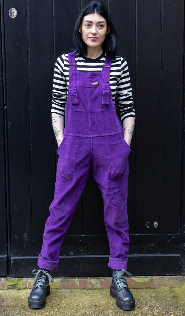 Gabrielle is wearing the purple magic stretch corduroy dungarees, paired with black and white striped long sleeve top and chunky boots. Model is standing against a black door, looking off camera with hands in pockets.