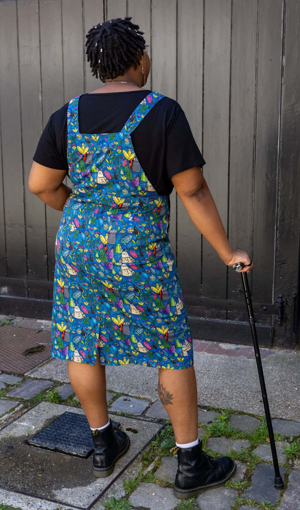 Han, a black femme model with silver glasses with a chain, is stood outside in Hove in a mews wearing Run & Fly x Katie Abey Crystal Critters Stretch Twill Long Pinafore Dress with a black t shirt underneath and black boots. The blue pinafore dress has an all over print of critters and crystals illustrated by Katie Abey. Han is stood with her back to the camera and is holding her black walking aid. 