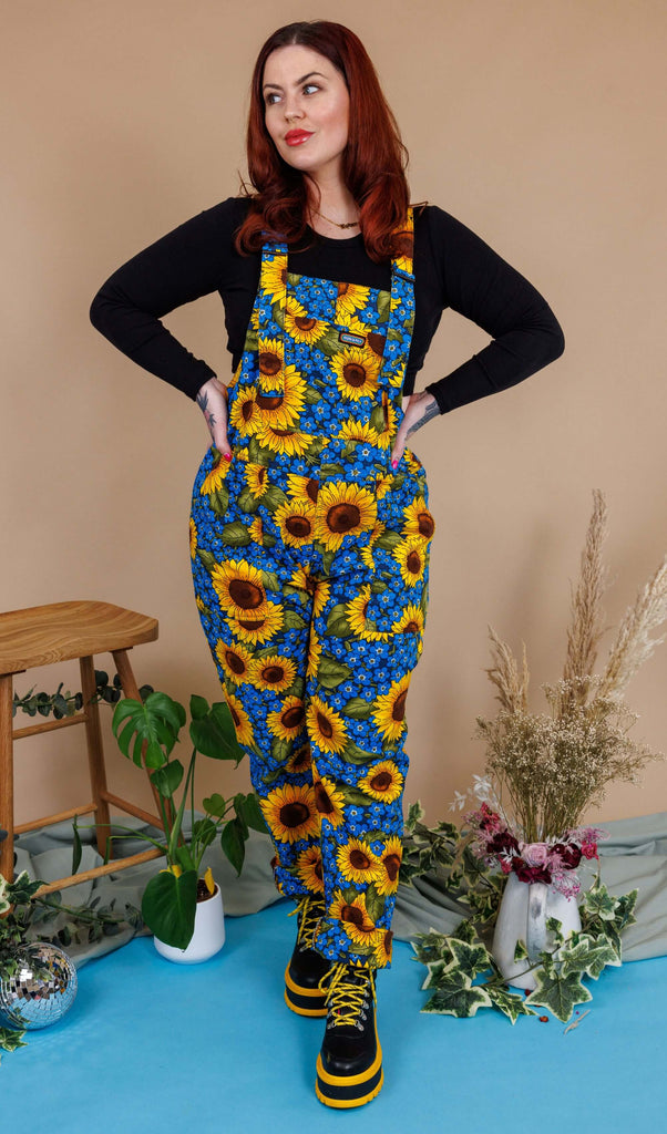 Aemelia Fox is stood in a photography studio in Hove with a tan colour backdrop and plants behind her wearing Run & Fly x Jen James Design Forget Me Not Charity Stretch Twill Dungarees with a long sleeve black top underneath and boots. The dungarees have an all over blue forget me not and yellow sunflower print. Aemelia is posing toward the camera with her hands on her hip and one leg in front of the other. She is looking off to one side.