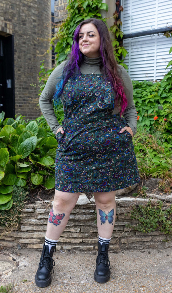 Luisa Christie wears the forest green paisley stretch twill pinafore dress, paired with green high neck long sleeve top and chunky black boots. Model is posing with hands in pockets, and looking away from the camera, in front of some greenery.