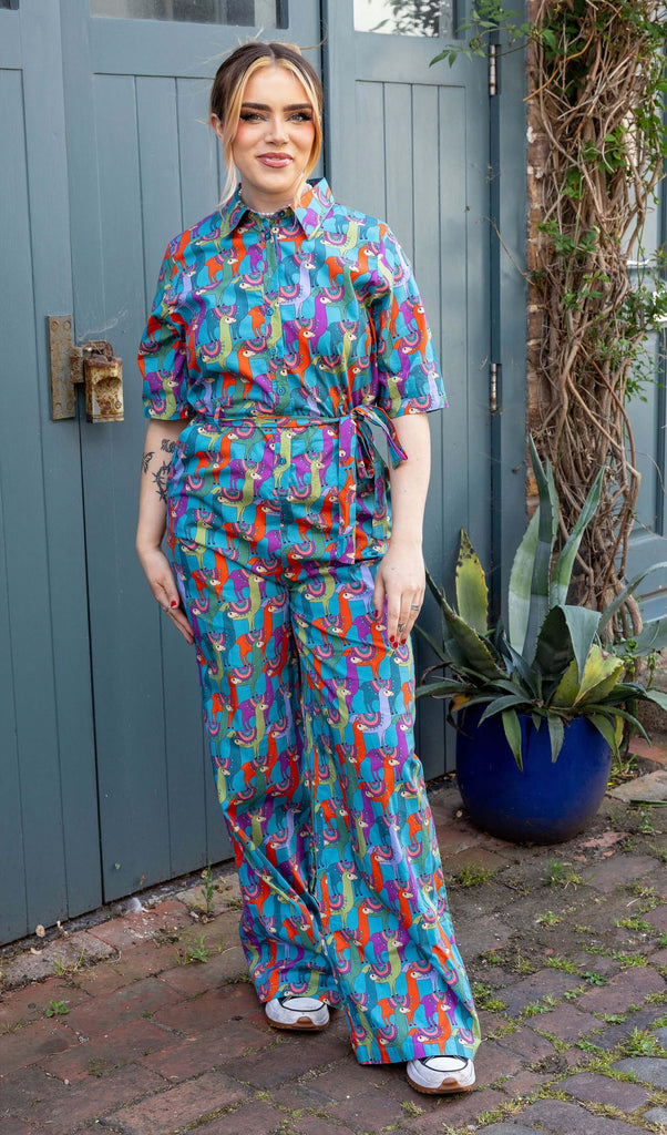 Model is wearing Party Llama Stretch Jumpsuit paired with trainers. The jumpsuit is a bright blue with llamas printed on in red, green, blue and purple. The jumpsuit is buttoned up to the collar and has a matching fabric belt tied round the front of the waist. Model is smiling at the camera.