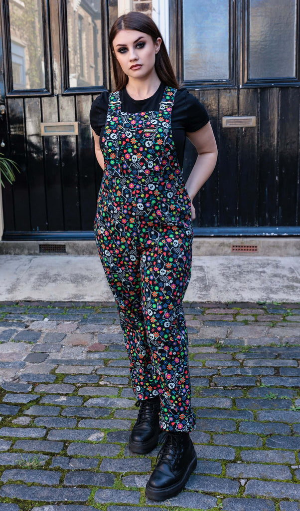 Charlotte is stood outside wearing the Halloween: Boogie Bones Stretch Twill Dungarees with a short sleeve black tshirt underneath and black boots. She is facing forward with both hands on her hips. Charlotte has long brown hair and dark smokey makeup. Boogie bones print is a black base with multicoloured flowers and dancing smiling cartoon skeletons all over.