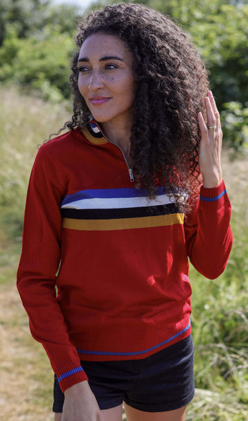 Rochelle is stood in a grass field wearing the red retro zip neck jumper with black shorts. They are facing forward posing with one hand running through their hair and the other by their side whilst smiling to the left. Photo is cropped from the hips up.