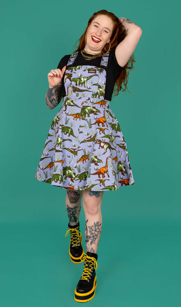 Izzy a red haired white model with tattoos is smiling wearing a blue dinosaur pinafore, black tee and black and yellow boots shot in studio against a green background